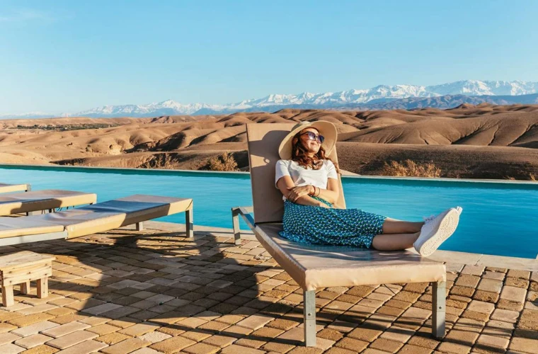 Woman relaxing on lounge chair at luxury desert camp pool overlooking golden Agafay stone desert and snow-capped Atlas Mountains
