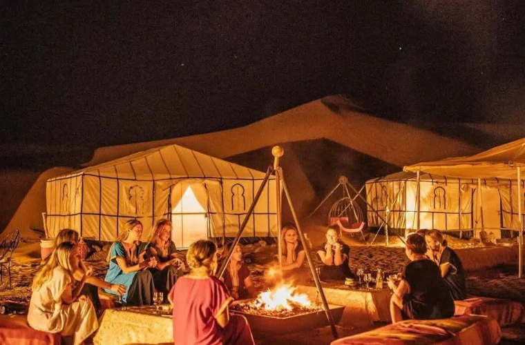 Tourists gathered around campfire at night in Sahara desert camp with illuminated traditional Berber tents and starlit sky